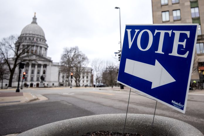A blue yard sign says "VOTE" in white letters with an arrow. The Wisconsin state capitol can be seen in the background.